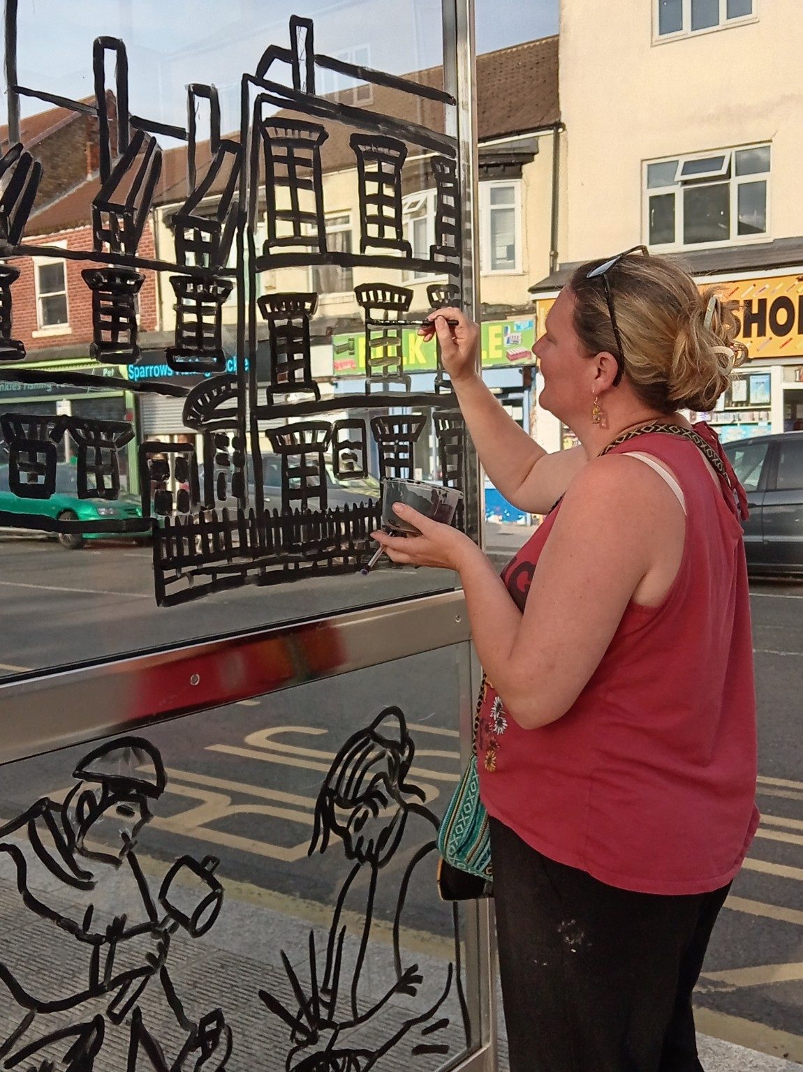 A person painting black street-scene outlines directly onto a bus shelter glass panel