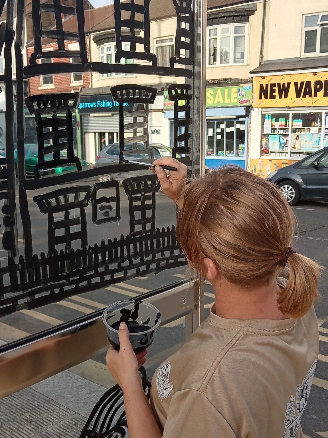 A person adding paint to a street-scene mural on a bus shelter with the road visible behind